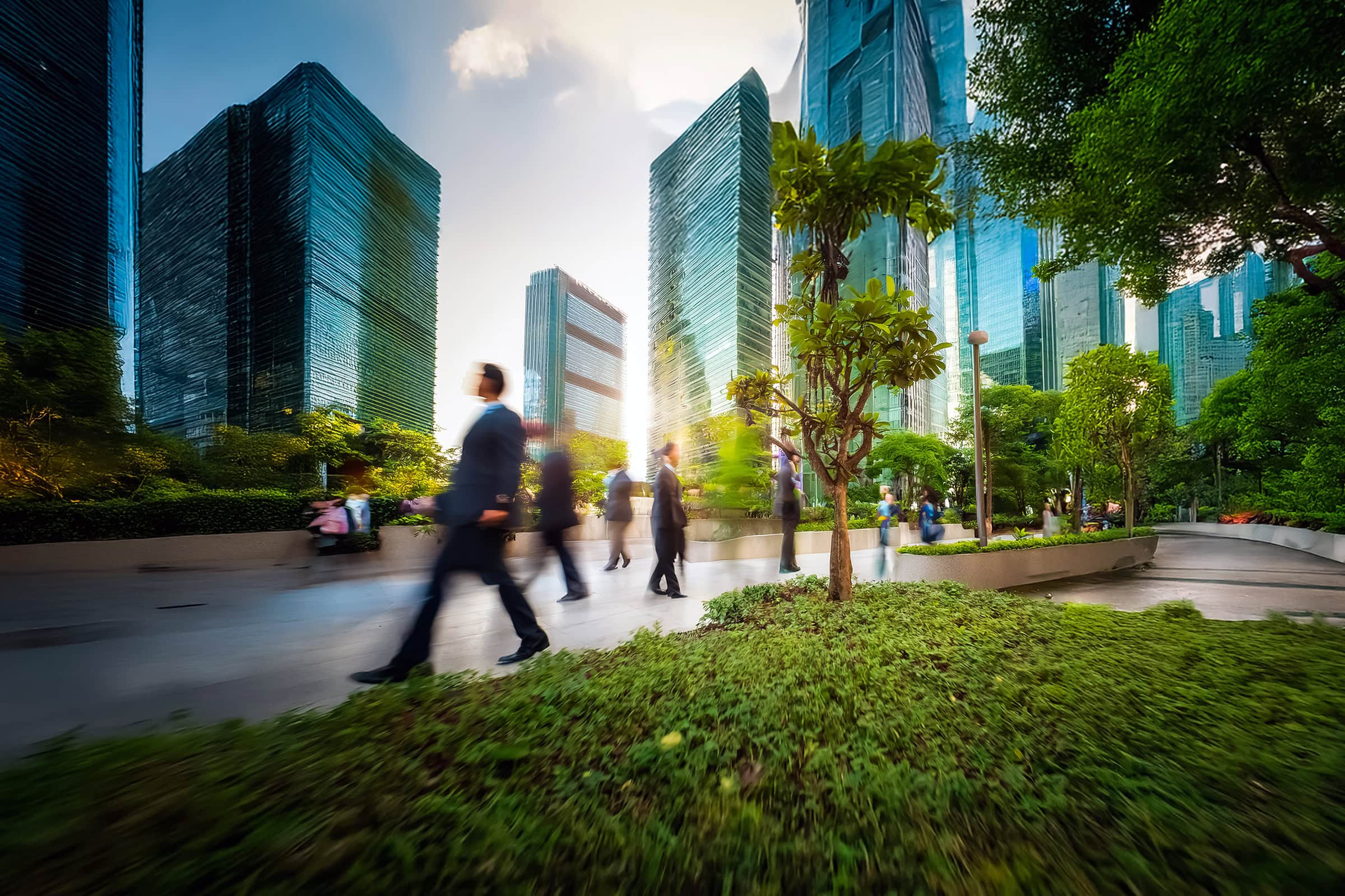 blurred-figure-walking-in-a-park Blurred figures walk in a park amidst towering skyscrapers. Lush greenery and trees provide a natural contrast to the modern urban architecture.