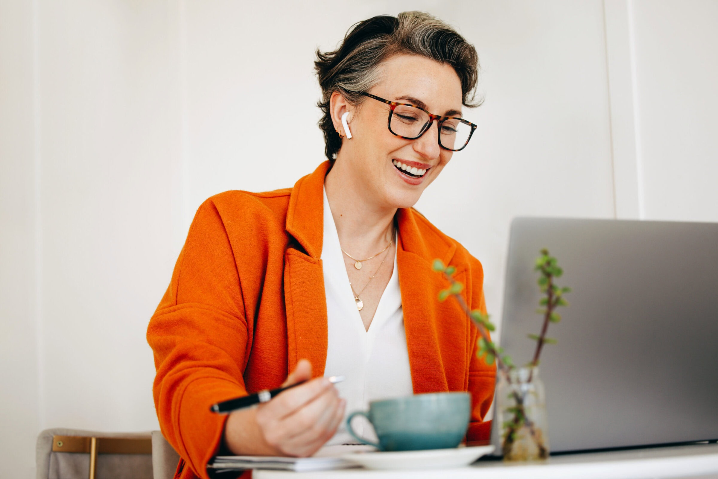 Smiling business woman waring an orange jacket attending a virtual meeting while working in a cafe.