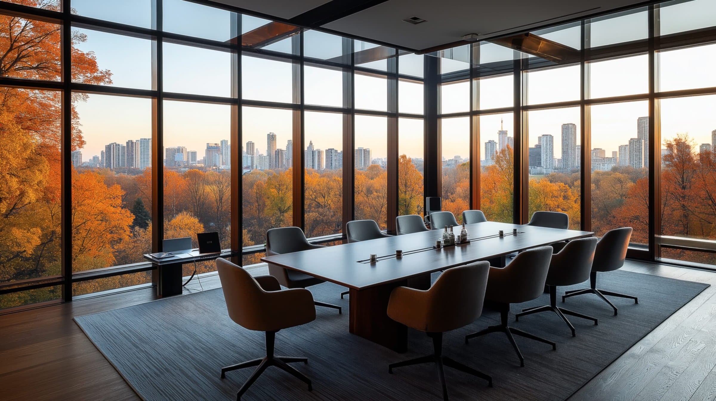 Modern conference room with large glass windows, overlooking a city skyline and vibrant autumn trees, with a long table and several chairs arranged around it.