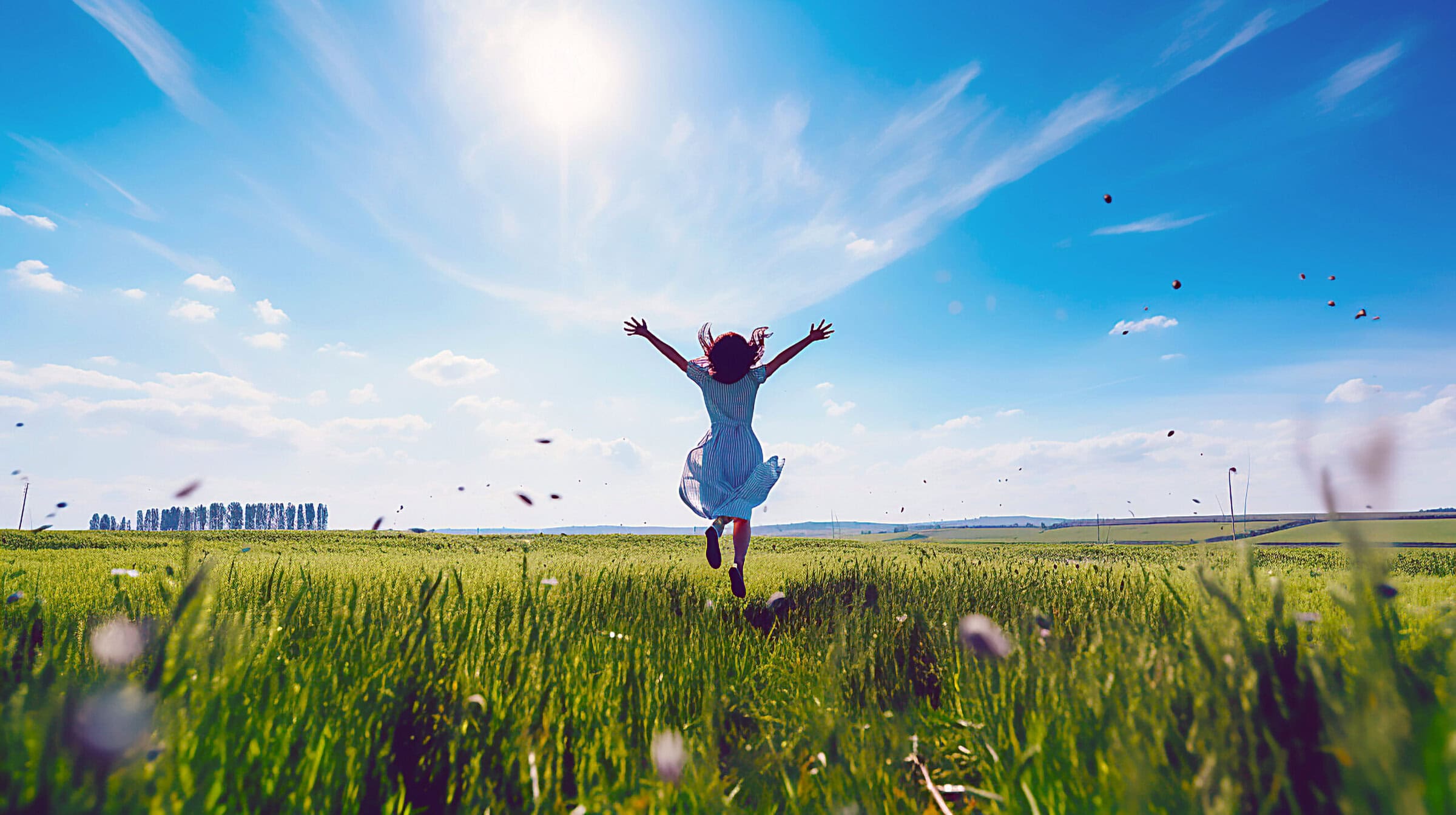 A woman jumps across a sunny meadow with her arms outstretched in joy.