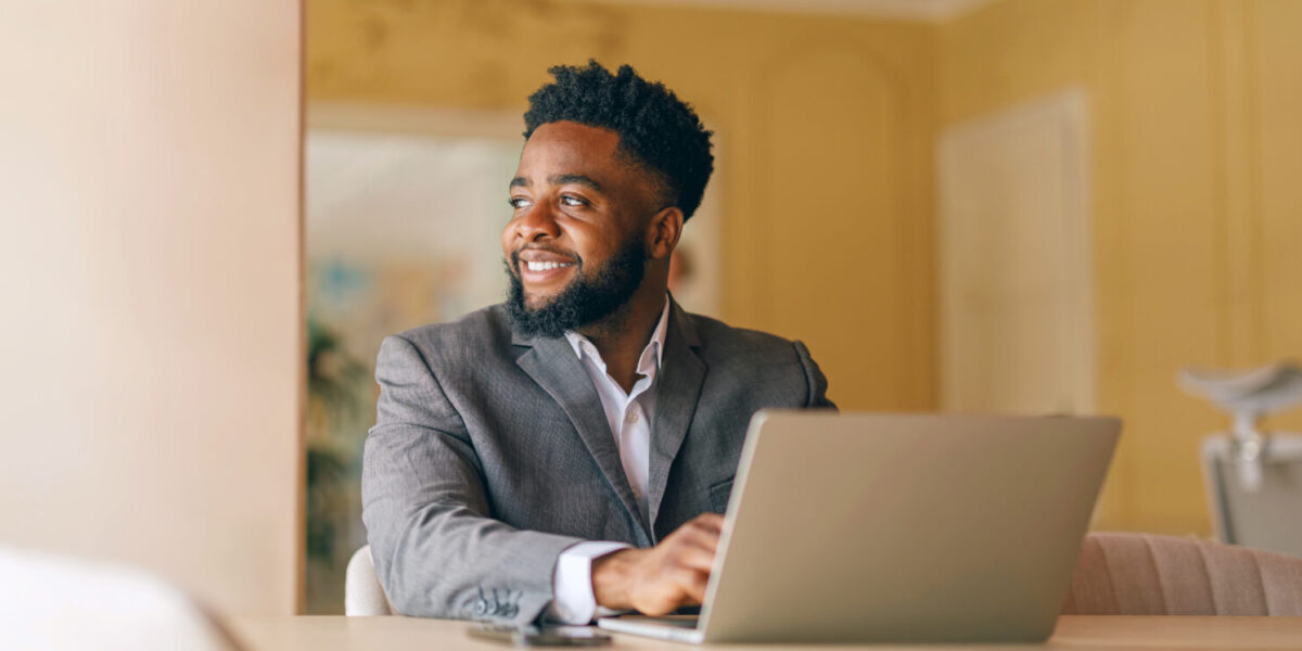 Male manager looking away and thinking business ideas while working over laptop at desk by wall in office