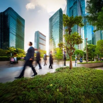 Blurred figures walk in a park amidst towering skyscrapers. Lush greenery and trees provide a natural contrast to the modern urban architecture.