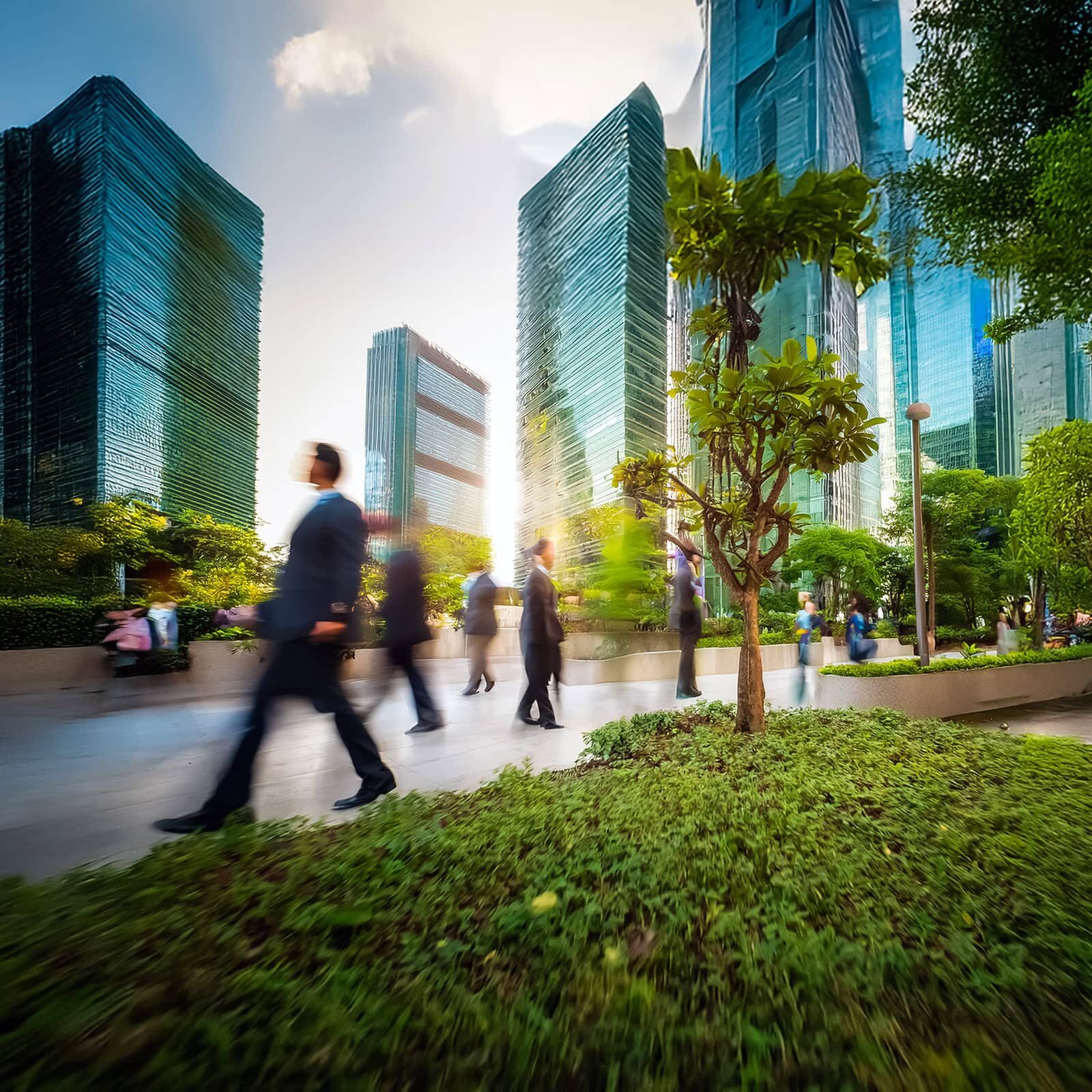 Blurred figures walk in a park amidst towering skyscrapers. Lush greenery and trees provide a natural contrast to the modern urban architecture.